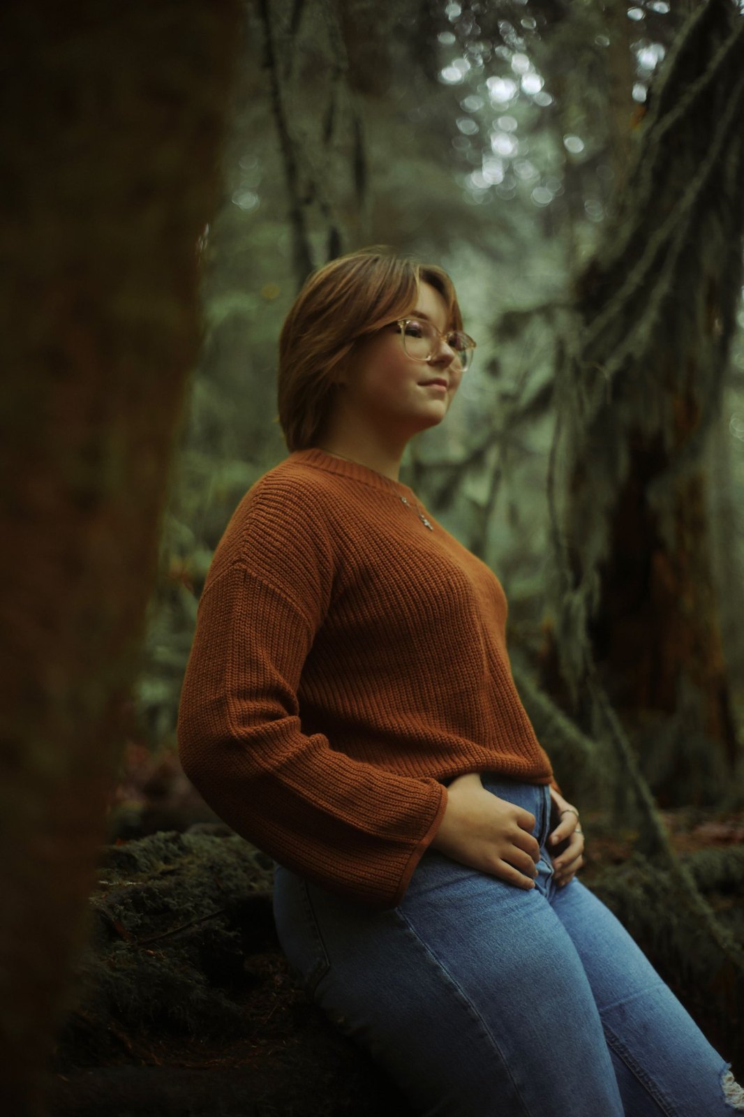 Senior in a rust sweater leaning back in a mossy forest, eyes on the light