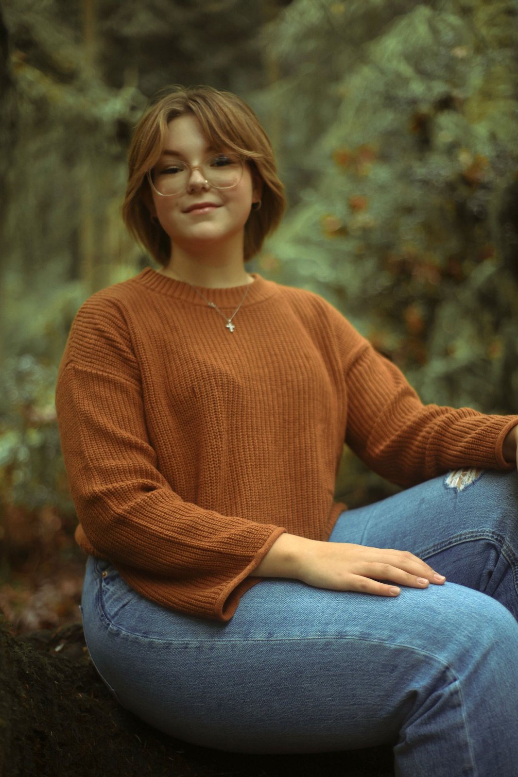 Senior in glasses and a rust sweater seated in the forest, smiling