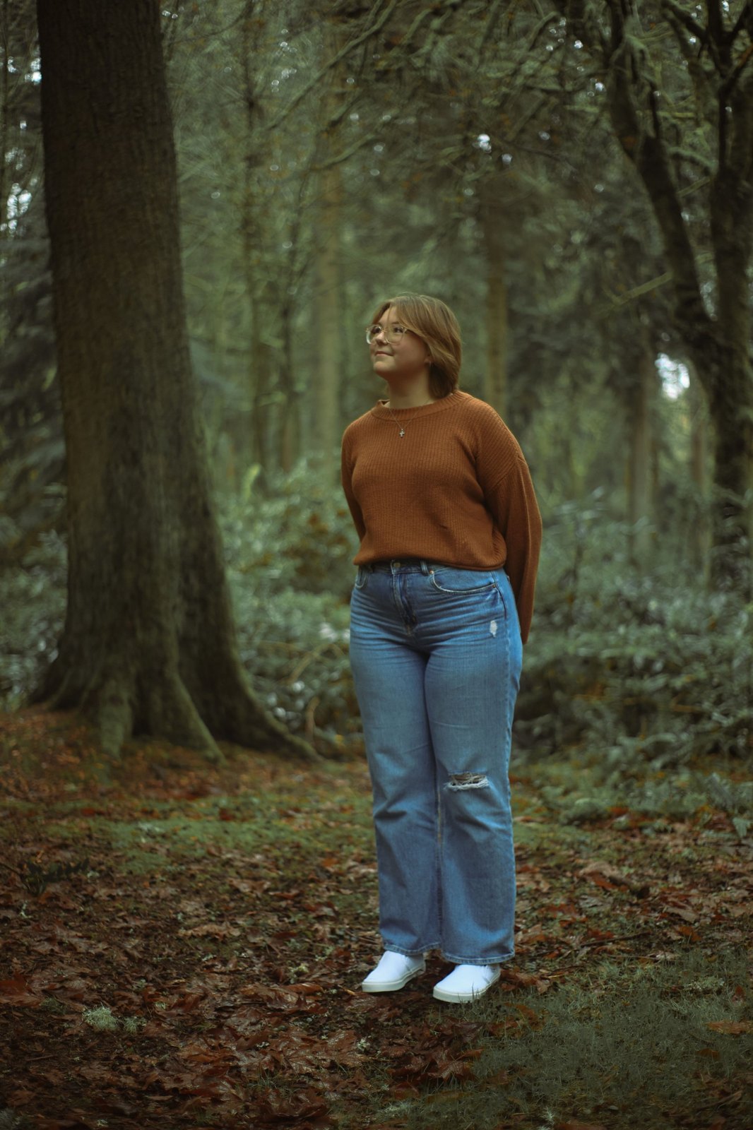 Senior in a rust sweater and jeans standing among tall forest trees